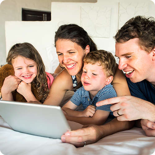 Parents lay on a bed with their son and daughter, laughing as they look over a tablet for affordable school photos that parents love.