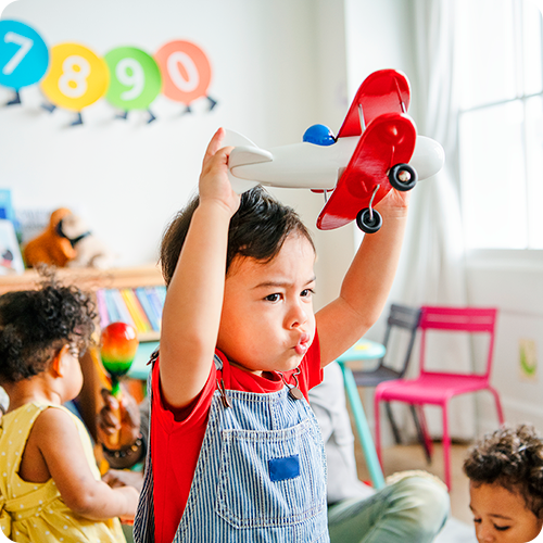 A young boy wearing a red shirt and blue overalls plays with a toy plane above his head in a kindergarten setting with children playing behind him.