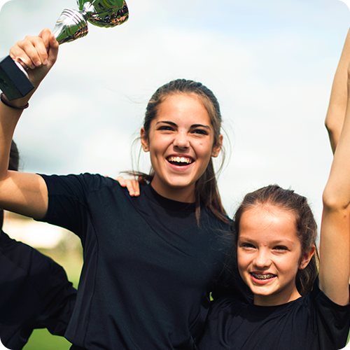 Two young, female athletes in black t-shirts cheering as one of them holds up a trophy.