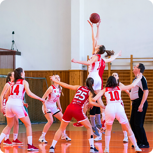 A sports photo of two girl's basketball teams competing in a gym with a referee present. The players are in a circle with two girls from opposing teams jumping to get a hold of the ball.