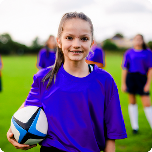 A sports photo of a young girl in a blue jersey smiling while holding a rugby ball, her teammates standing behind her blurred out.