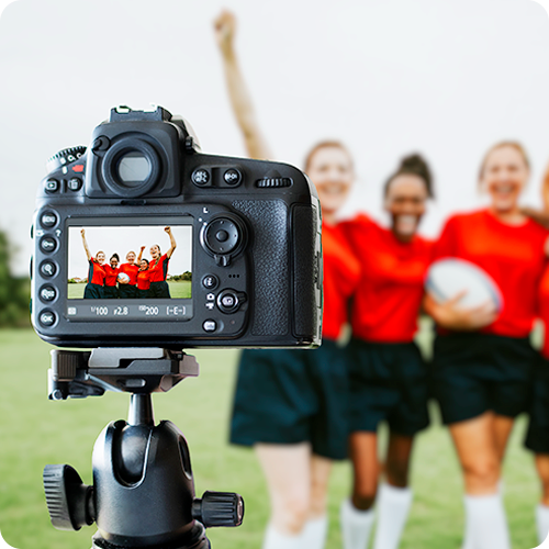 A sports photography image of a camera in the foreground photographing a rugby team in red jerseys. The rugby team is shown behind the camera at a distance, cheering and out of focus.